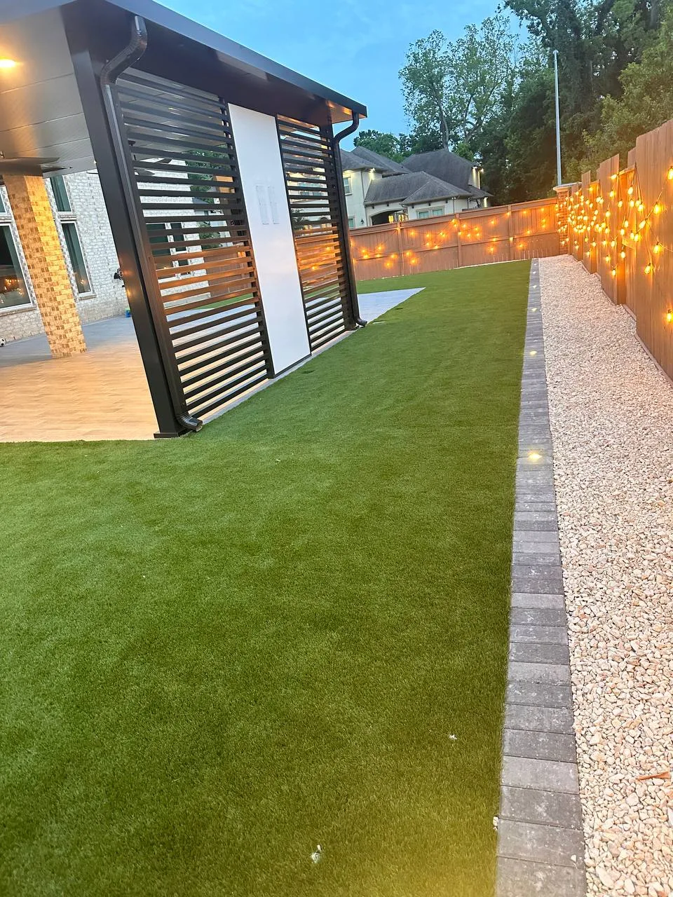 Modern aluminum louvered pergola with artificial turf, gravel path and string lights at dusk in Houston TX
